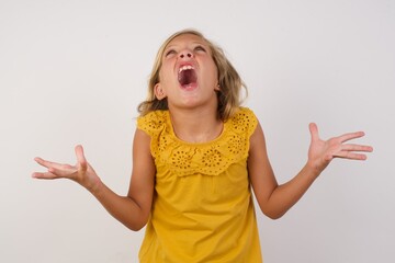 Young blonde kid girl wearing yellow dress over white background crazy and mad shouting and yelling...