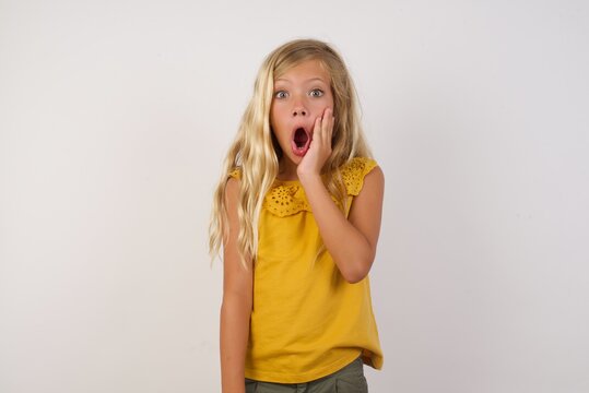 Horizontal portrait of shocked Young blonde kid girl wearing yellow dress over white, looks with great surprisment being very stunned, astonished with unexpected news, . Facial expressions concept.