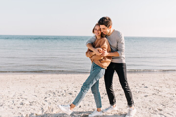 Emotional brunette guy posing at sea beach with girlfriend. Outdoor portrait of smiling couple enjoying date at ocean.