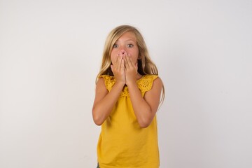 Young blonde kid girl wearing yellow dress over white background or studio wall, giggles joyfully, covers mouth, has fun alone,  has natural laughter, hears positive story or funny anecdote