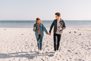 Full-length portrait of couple drinking coffee at ocean beach. Well-dressed guy holding hands with girl on sea background.