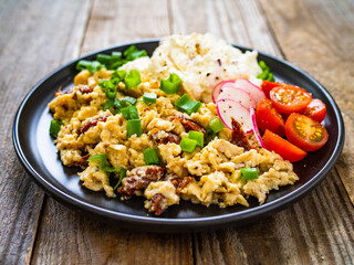 Delicious breakfast - scrambled eggs with fried sausages, cottage cheese and vegetables served on black plate on wooden table
