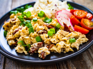 Delicious breakfast - scrambled eggs with fried sausages, cottage cheese and vegetables served on black plate on wooden table
