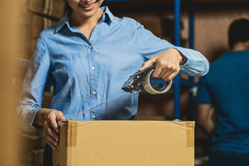 Closeup of Warehouse worker woman packing cardboard box with sticky tape with Indian worker man in...
