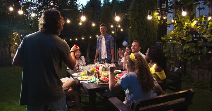 Happy multi-ethnic people having fun and playing at garden party in evening. Caucasian boys and girls drinking and talking. African American and Asian females laughing. Party games. Fun concept