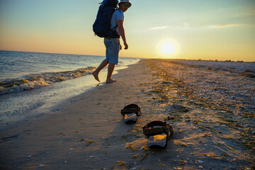 sandals on wet sand near the sea. The guy walks with a backpack along the coast. The rays of the...