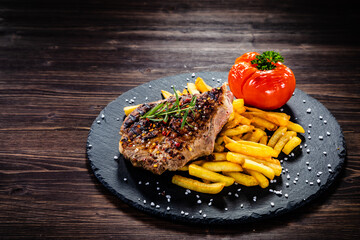 Grilled steak with french fries and vegetables served on black stone on wooden table
