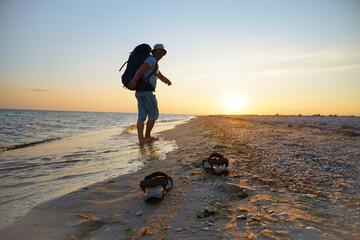 sandals on wet sand near the sea. The guy walks with a backpack along the coast. The rays of the...
