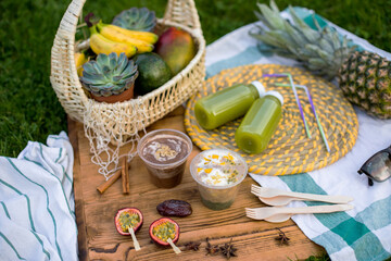 Vegetarian picnic. Desserts from tropical fruits mango, pineapple, banana and maracuu stand on a wooden background. Next to it are two green smoothie bottles and a basket of tropical fruits. Diet food