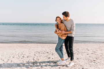 Ecstatic girl in jeans standing on sea background. Outdoor portrait of happy couple posing at sandy beach.