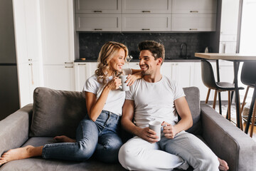 Couple in love sitting on sofa holding cups looking at each other and smiling. Romantic couple enjoys morning together at home.