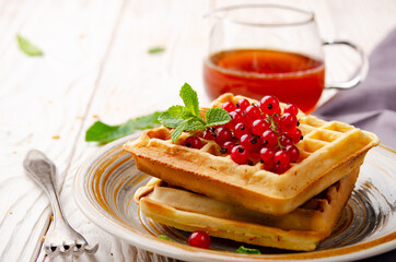 Belgian waffles served with red currants and mint leaf on white wooden kitchen table with syrup aside