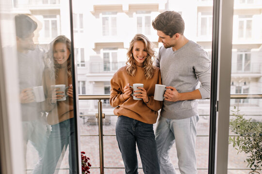 Blissful man standing at balcony with wife. Portrait of cute couple enjoying coffee.