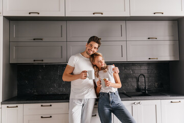 Smiling european man embracing wife in kitchen. Indoor photo of pretty girl drinking coffee in weekend morning.