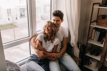 Happy guy in jeans sitting at home with wife. Smiling blonde girl chilling with boyfriend in...