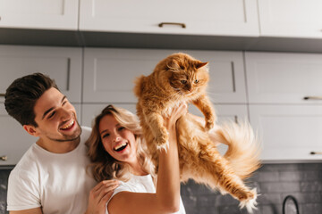 Excited couple posing with fluffy cat. Indoor portrait of smiling adorable woman holding her pet in kitchen.