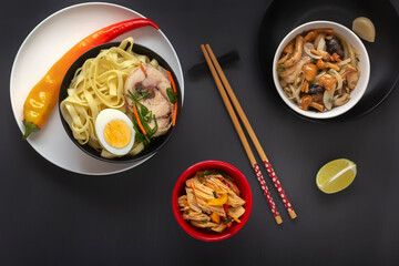 Noodles with pieces of meat and egg in a black plate and dim sums with different traditional snacks of mushrooms, soy and vegetables with chopsticks on a dark table. Top view. Asian food concept