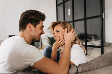 Romantic smiling couple in love sitting on sofa at home and looking at each other. Tender man with his beautiful girlfriend enjoying morning together.