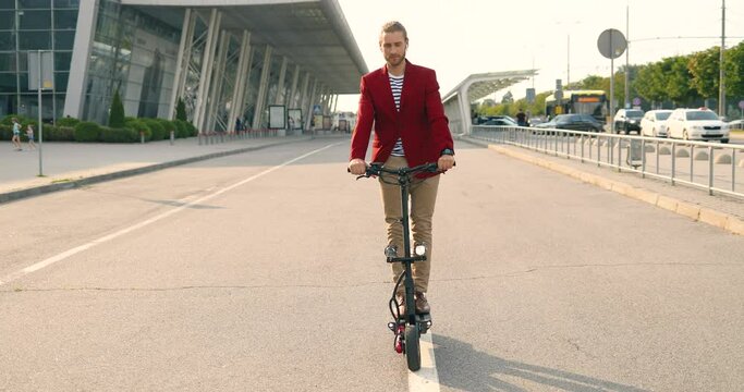 Handsome Caucasian young stylish man in red jacket standing on electric scooter at urban street near big modern building. Male rider going by city on the road. Sunny day.