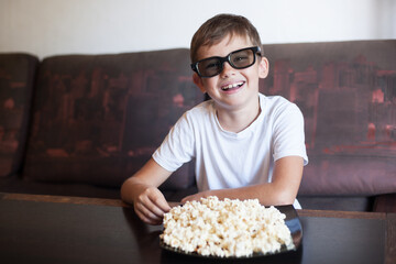 a boy in 3D glasses watches a movie and eats popcorn smiling broadly sitting on the sofa at home