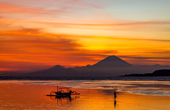 Small traditional boat silhouetted against a volcano at sunset with reflection