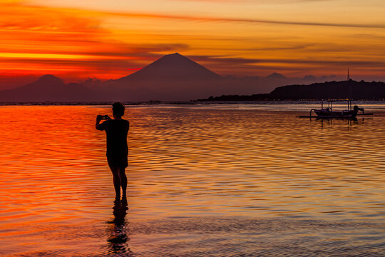 Silhouette of a man taking a photo of a volcano and traditional boats at sunset