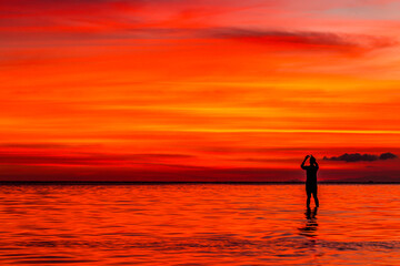 Fototapeta premium A person stands in the water silhouetted against the sun, taking photos of the sunset