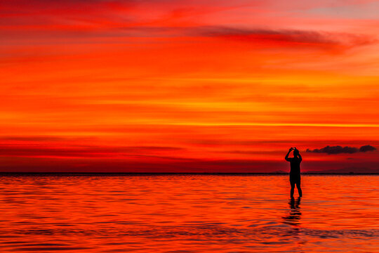A person stands in the water silhouetted against the sun, taking photos of the sunset