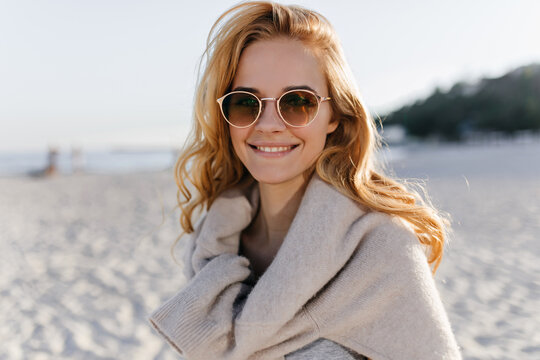 Blonde curly girl tied cashmere sweater around neck and looking at camera with smile, posing in glasses on beach