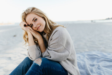 Green-eyed woman in jeans and sweater is sitting on sand, leaning on her knees and looking into camera against background of sea