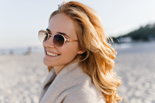 Close-up portrait of positive girl with wavy blonde hair dressed in beige cashmere sweater and sunglasses on beach