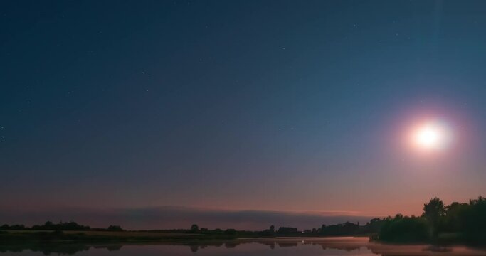 Starry night sky above a calm lake with flat water surface. Stars and moon timelapse