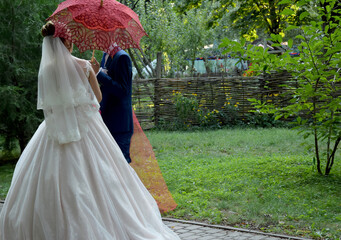 Fototapeta premium The bride and groom are walking in the park under a red lace umbrella.