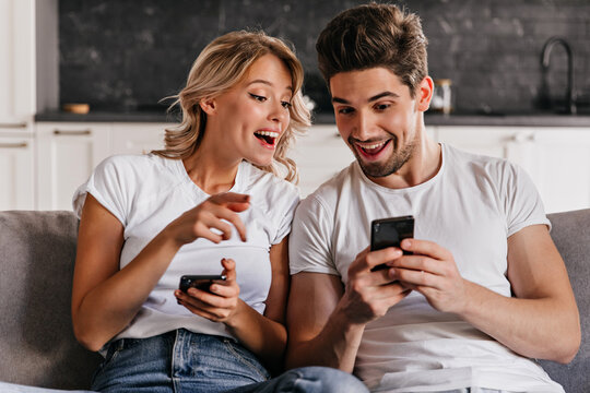 Smiling couple sitting on couch with phones. Adorable young woman holding smartphone.