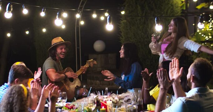 Joyful multi-ethnic young friends having fun at party in garden at summer evening and applaud. Caucasian guy playing the guitar and singing outdoors. African American and Asian girls smiling.