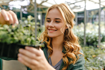 Long-haired redhead girl is happy and sincerely smiling, holding pot of greens in her hands. Closeup portrait outside surrounded by plants