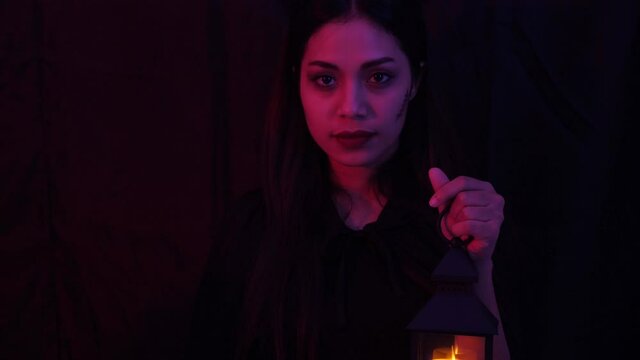 Close up, portrait of young pretty and sexy Asian woman in halloween concept costume, holding lantern, staring to camera, smile and beckoning over dark background with red and perple light setting