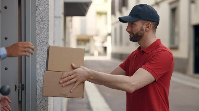 An young smiling mailman courier in uniform is delivering a parcel directly to a customer home. Concept of courier, delivery, e-commerce, online shopping
