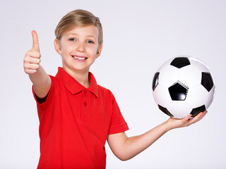 Photo of a smiling boy in sportswear holding soccer ball, posing at studio. Happy 8 years old kid in a red t-shirt with a soccer ball in hand. White child with a smile holds a soccer ball.