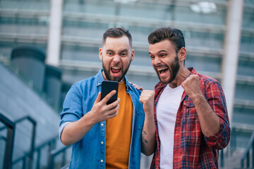 Two best friends outdoors. Guys excited and happy due to winning gamble with smartphone
