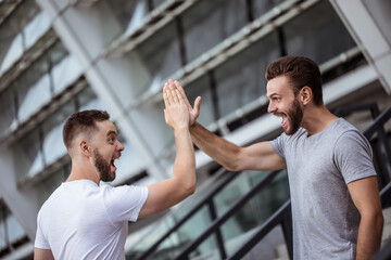 Give me five! Two happy excited modern bearded best friends in casual clothes having fun and chatting together outdoors.