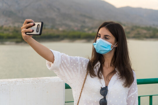 Happy girl in white with mask taking selfie photograph with her smartphone in a mountain lake.