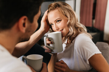 Beautiful blonde girl in love looking at her boyfriend and drinking coffee from cup. Tender cute photo of romantic couple at home.