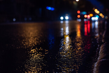 Fototapeta na wymiar The blurred focus of the city road and car during rain at night, with colorful unfocused lights on the rear background. Water from the rain and an abstract image of the side of the lights in the city
