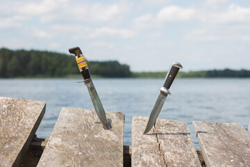 Two hunting knives stuck in the boards on the background of the lake