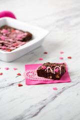 Obraz na płótnie Canvas Pan of Homemade Brownies with Pink Frosting on a White Countertop with Heart-Shaped Sprinkles; Piece on Pink Napkin and White Heart-Shaped Doily in Front with Heart-Shaped Sprinkles Scattered Around