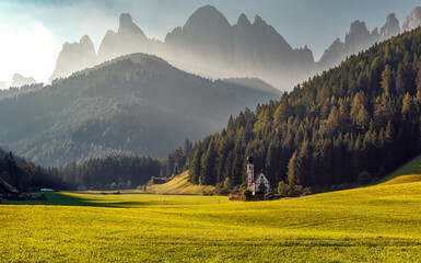 Fototapeta na wymiar Magic foggy morning scenery during sunrise over the Dolomites mountains. Fantastic summer View on mountain valley with green grass and famouse church in Santa Maddalena village. Italy