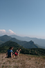 Fototapeta premium Dog traveler goes to the mountains with his best friend man and enjoys nature and beautiful scenery. A young traveller girl is sitting on top of a mountain with her German shepherd.