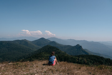 Fototapeta premium Beautiful mountain and hilly landscape in warm, Sunny weather and the young tourist. A female traveler sits on top of a hill and looks at the surrounding mountains.
