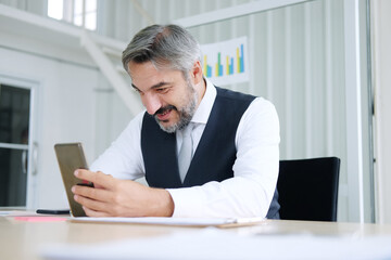 Naklejka premium Smiling smart Caucasian senior manager and businessman in black suit relaxing in social media online and talking, calling with businesspeople on smartphone at workspace in office